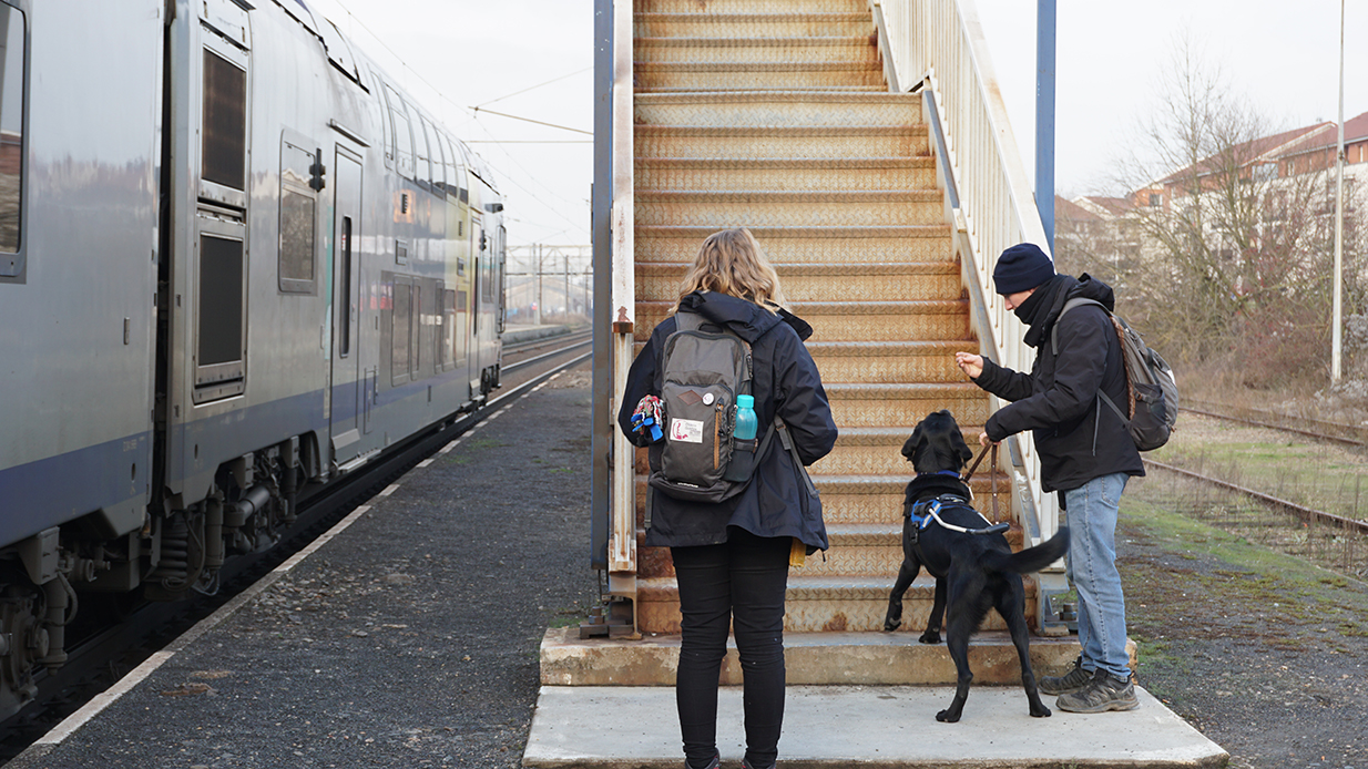 Homme et femme avec un chien noir devant un escalier sur un quai de train