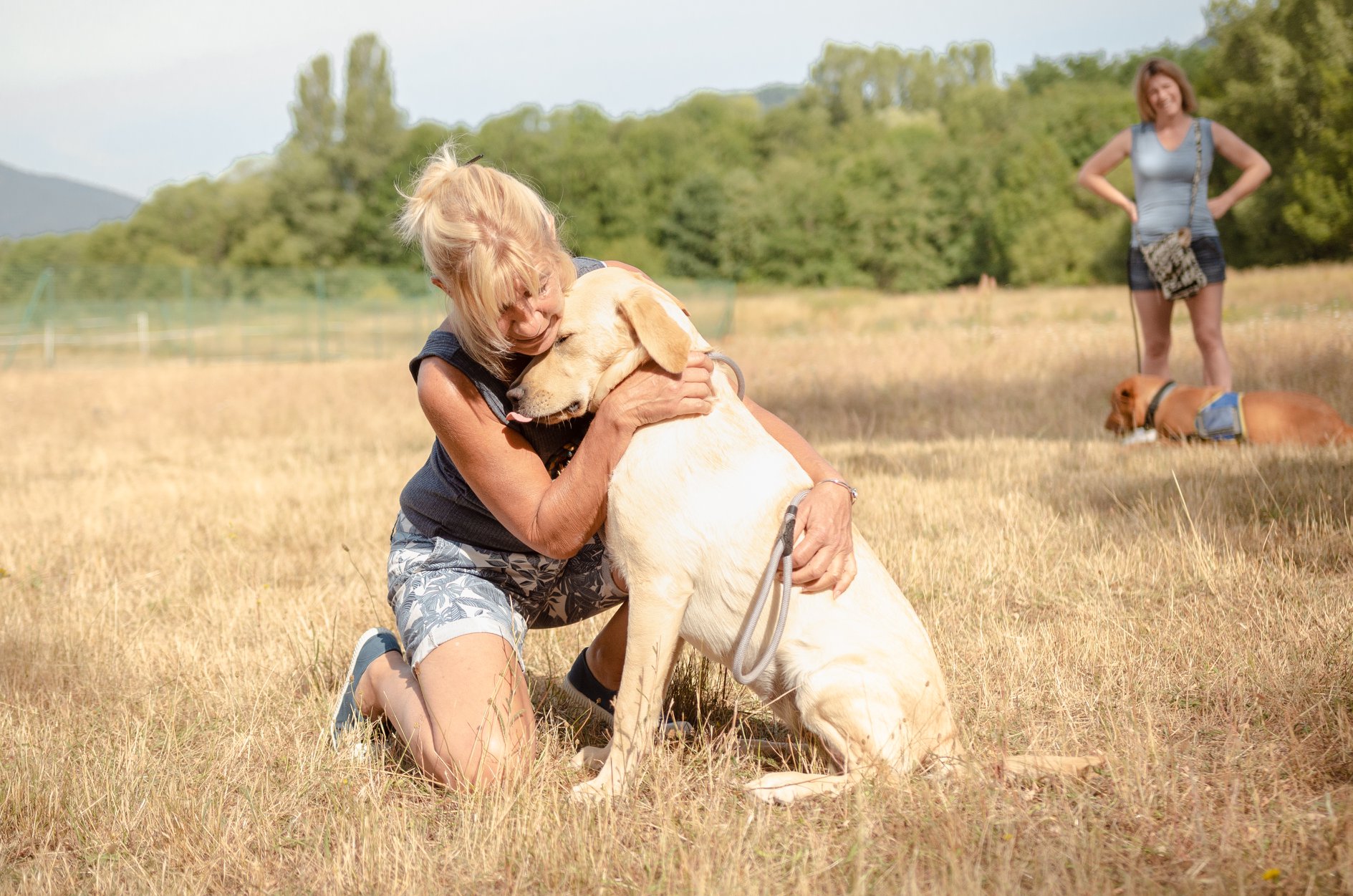 Femme accroupie faisant un calin à un chien avec une femme et un chien en fond